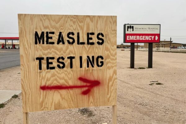 A wooden sign with “MEASLES TESTING” painted in black capital letters and a red spray-painted arrow pointing right, located outside Memorial Hospital in Seminole, Texas, with an “EMERGENCY” sign visible in the background.