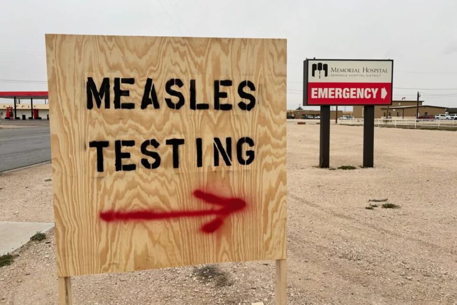 A wooden sign with “MEASLES TESTING” painted in black capital letters and a red spray-painted arrow pointing right, located outside Memorial Hospital in Seminole, Texas, with an “EMERGENCY” sign visible in the background.