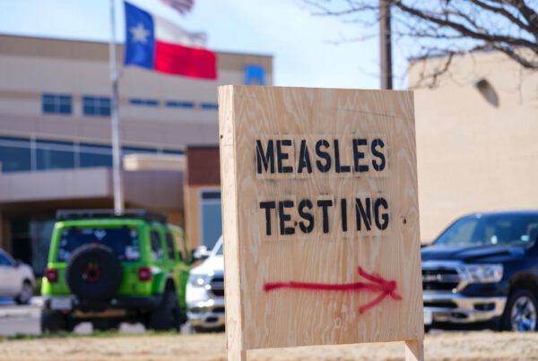 A wooden sign with bold black letters reading “MEASLES TESTING” and a red spray-painted arrow pointing right, positioned outside a medical facility with the Texas flag in the background.