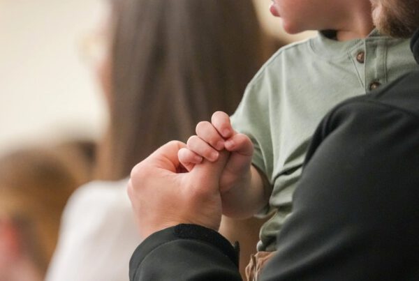 A close-up of a young child holding an adult’s thumb during a church gathering.